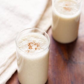 banana milkshake in two glasses with a light sprinkle of ground cinnamon on a dark brown table with a cream jute napkin by side