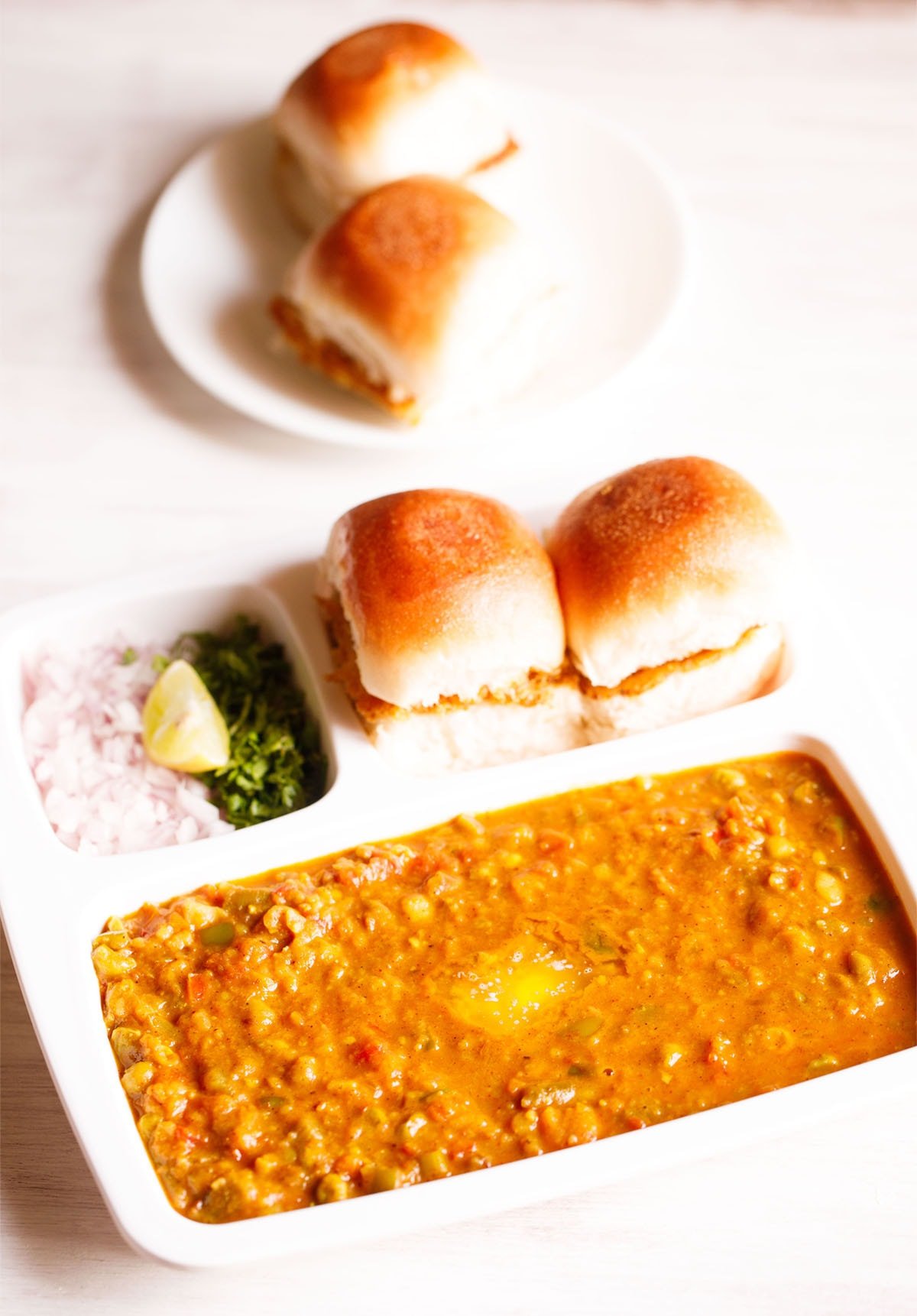 mumbai pav bhaji served in a rectangular serving tray with buttered pav and chopped onions, coriander leaves and lemon wedges on a white table