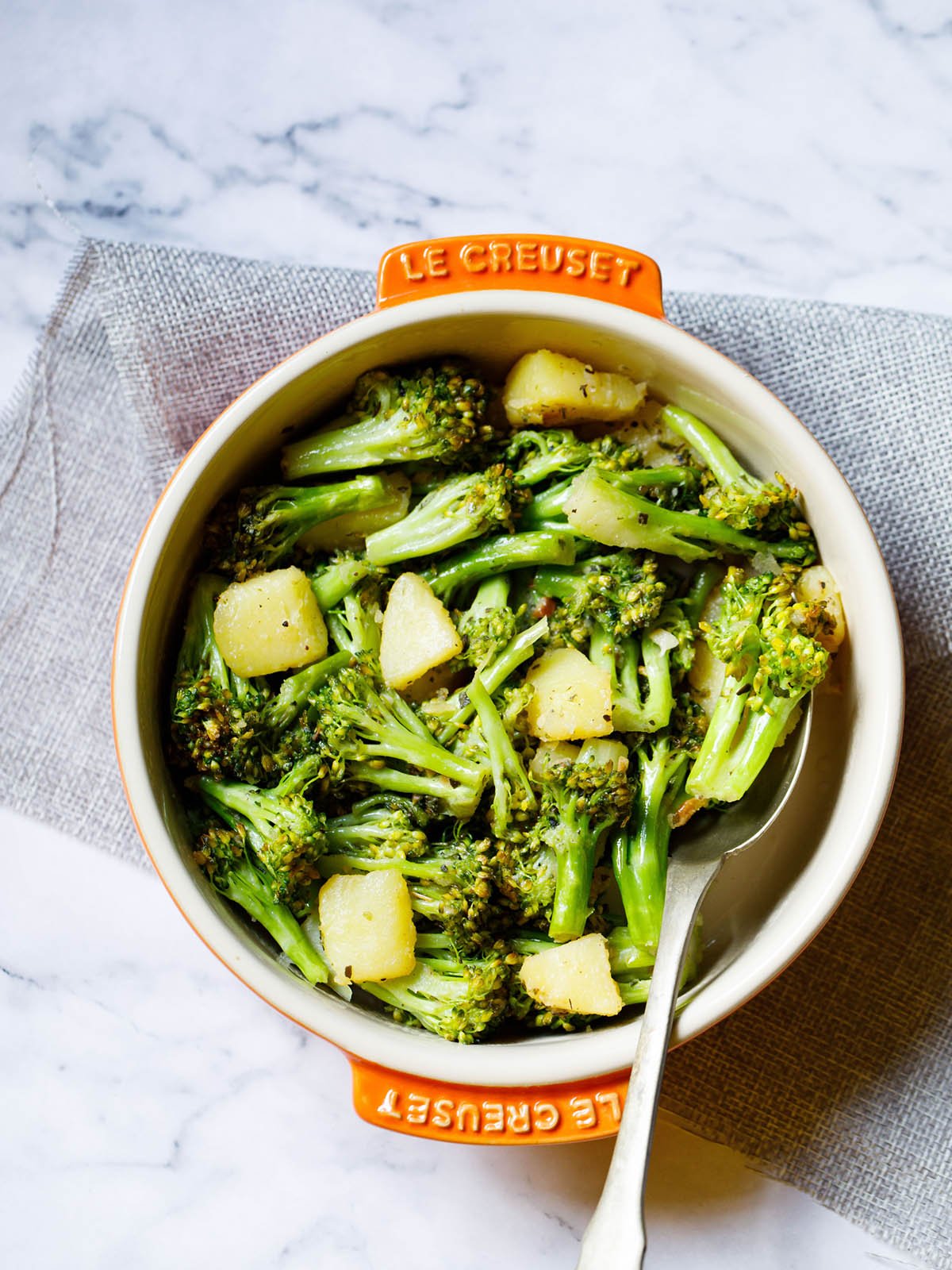 sauteed broccoli in a le creuset cream ceramic pan with orange handles with a spoon inside placed on a folded light gray jute fabric