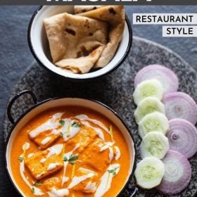 paneer butter masala served in a blue rimmed white pan, garnished with cream and cilantro with a side of roti, sliced cucumber and onions.