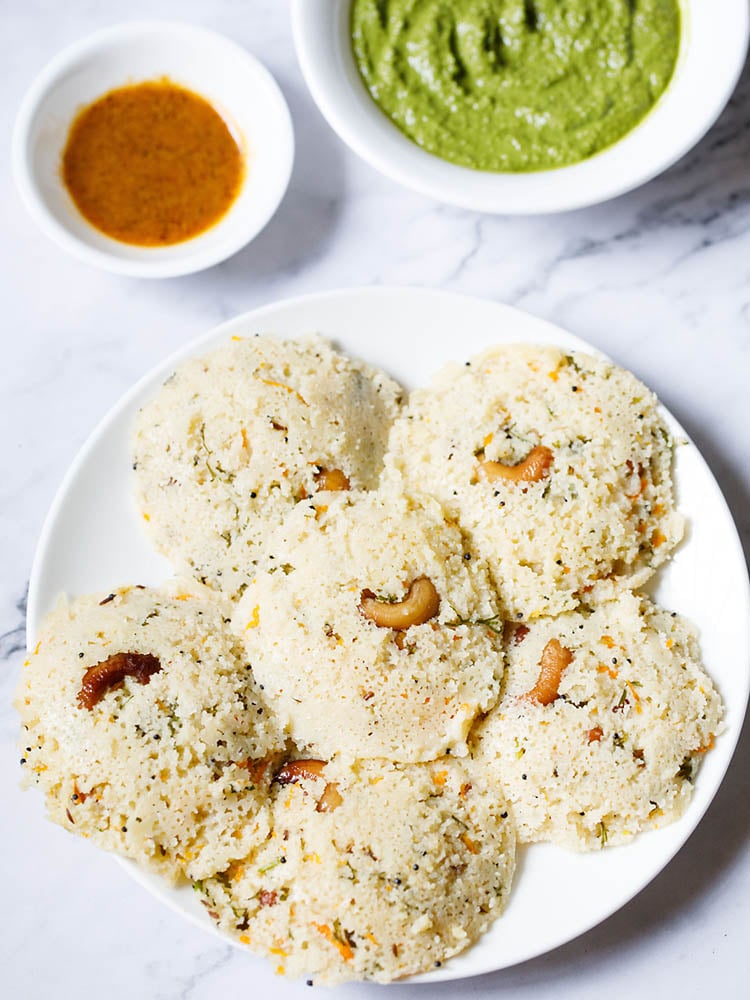 rava Idli arranged in a circular way on a white plate kept on a white marble backdrop with two chutney (dips) placed above the plate.